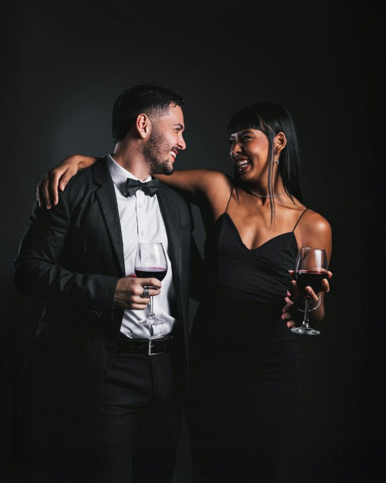 Couple dressed in formal evening wear enjoying red wine, smiling warmly in a dark, intimate setting.